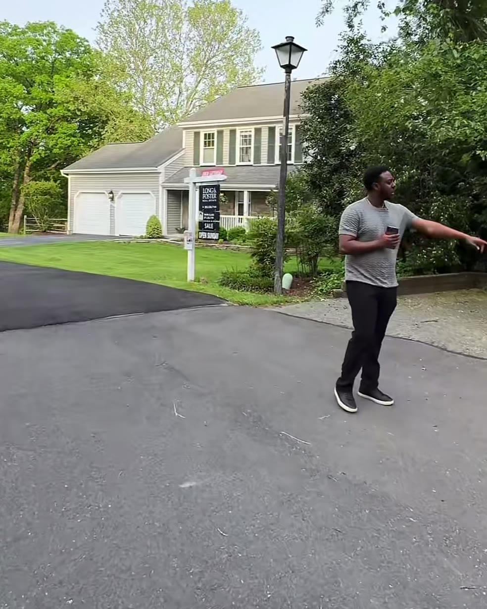 Adonay on the driveway pointing toward the house, with a lamp post and the listing sign