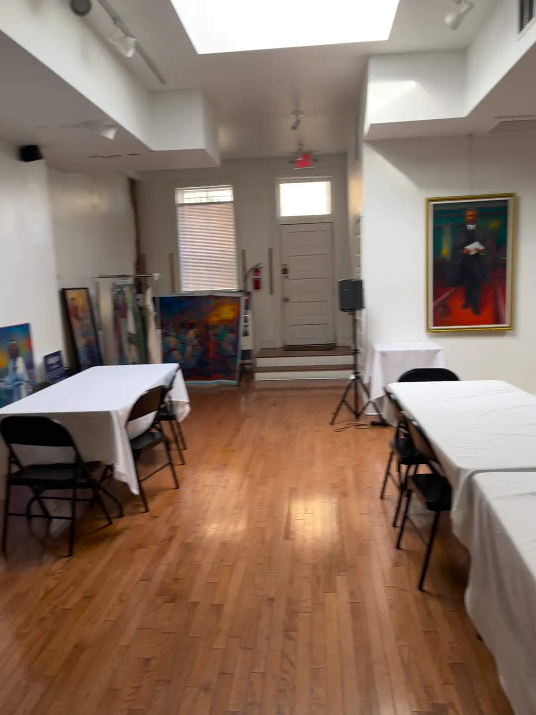 White-linen tables and folding chairs in the event space, with a portrait painting on the wall