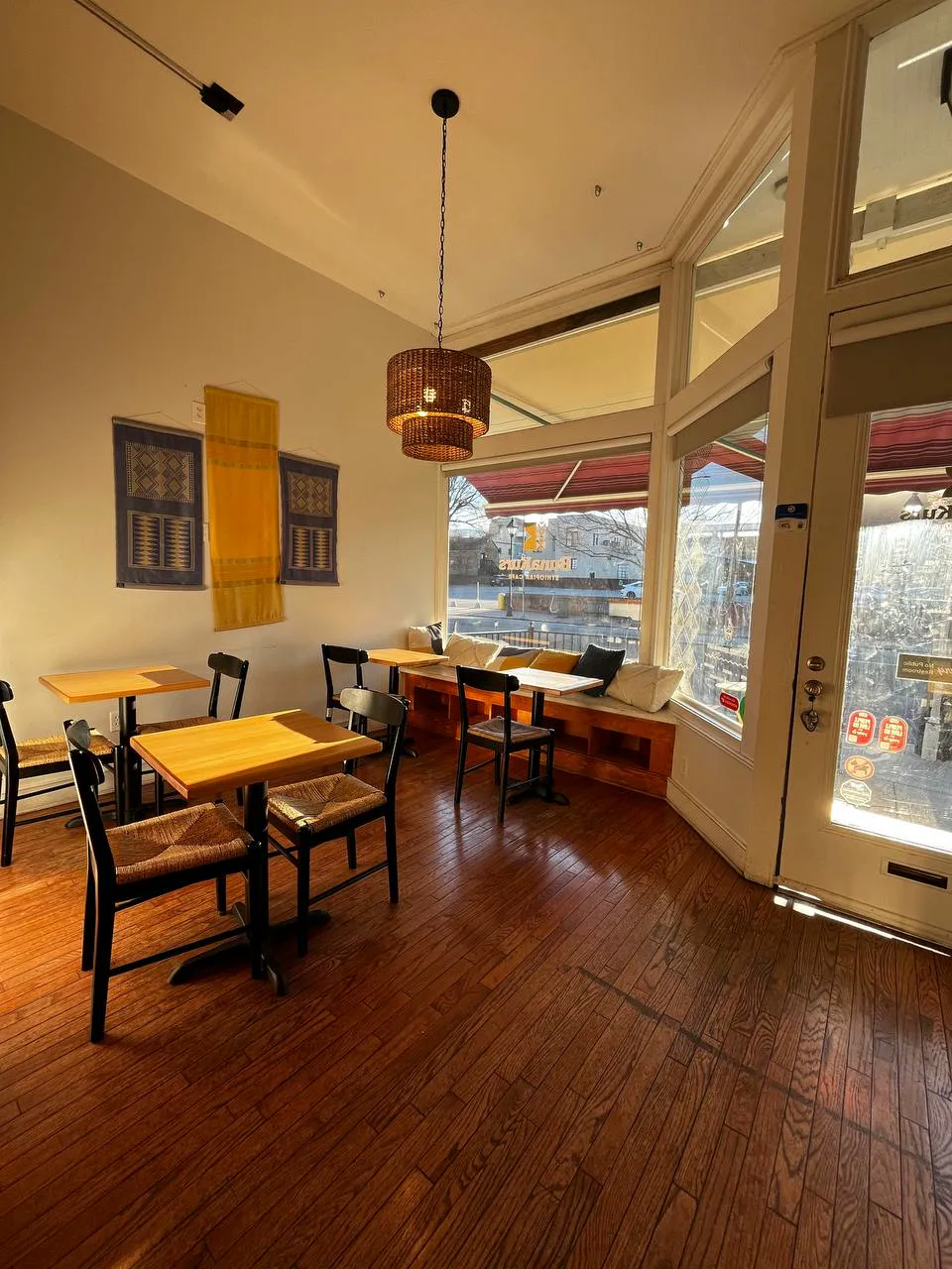 Sunlit corner with small wooden tables, rush-seat chairs and a cushioned window bench