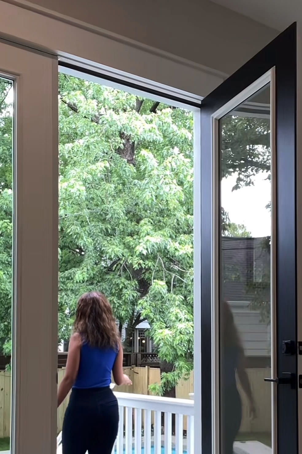 The agent in a cobalt top opening tall black-framed glass doors onto a deck with trees beyond