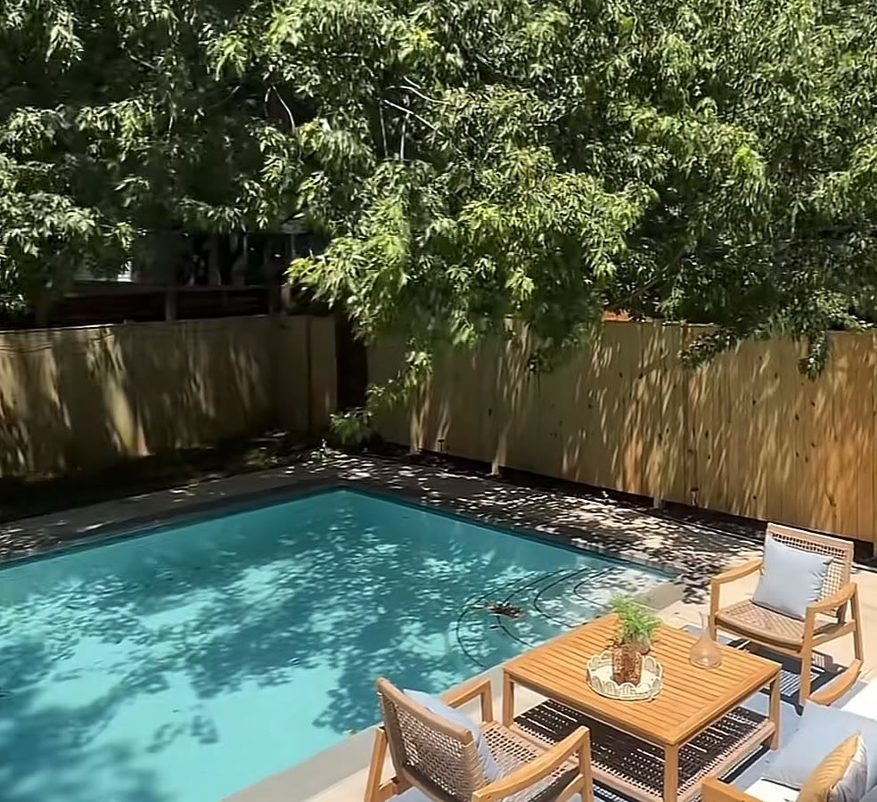 In-ground pool shaded by mature trees, with a teak lounge set on the patio and a tall wood privacy fence