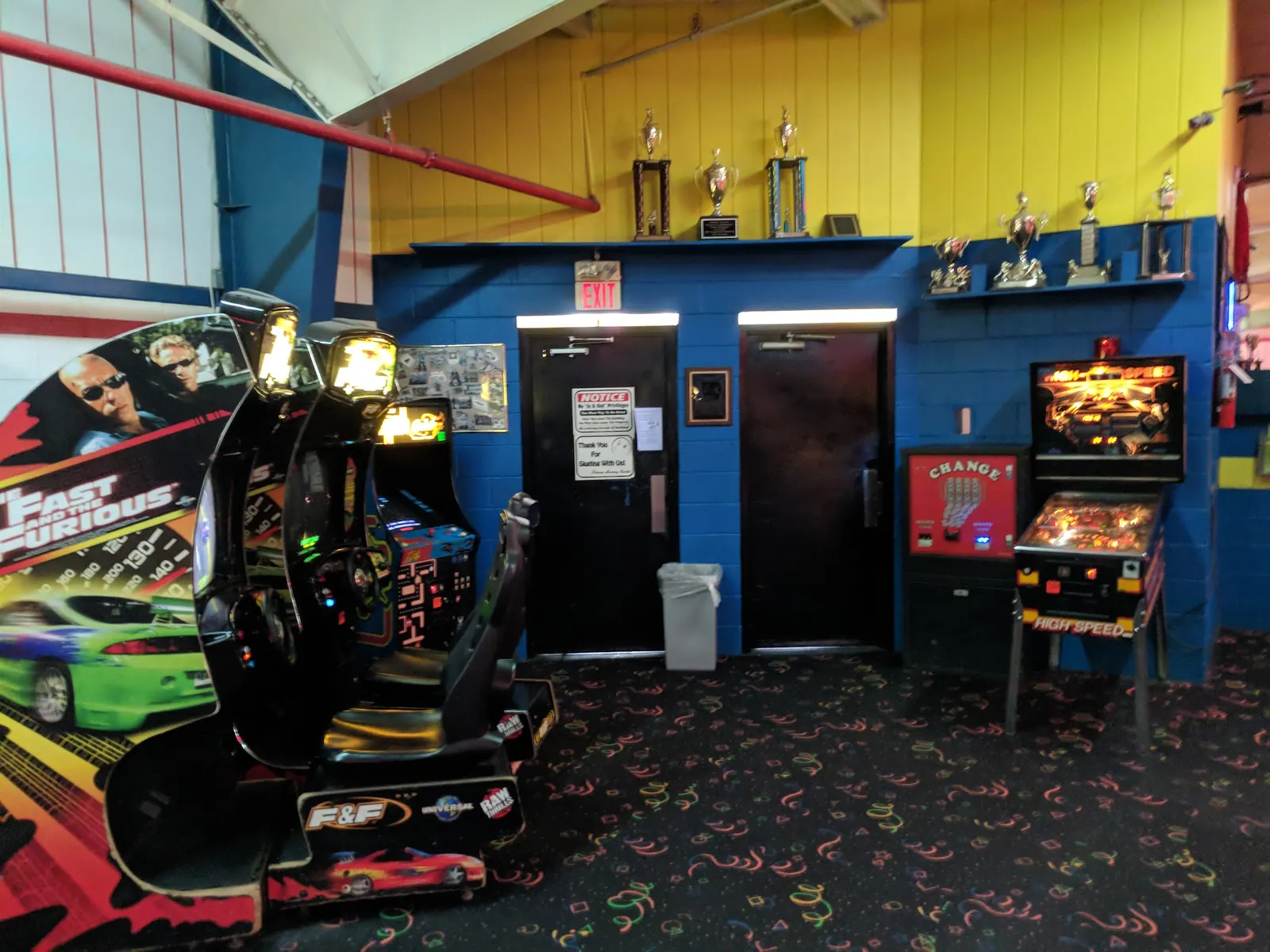 Arcade corner with a racing game and a pinball machine beneath a shelf of trophies.
